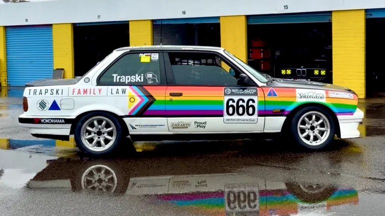A white BMW race car in Trapski Family Law livery with a progress pride stripe and the number 666, parked in front of pit garages